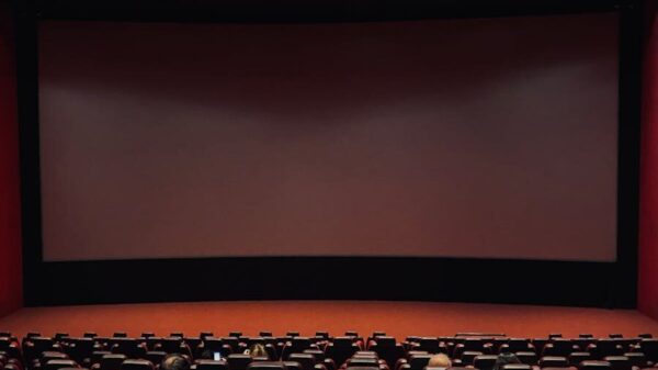 Dimly lit cinema hall with red seats and a blank screen. Minimal audience.
