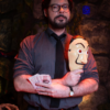 A bearded man stands in a dimly lit room holding a mask and money, exuding an air of mystery.
