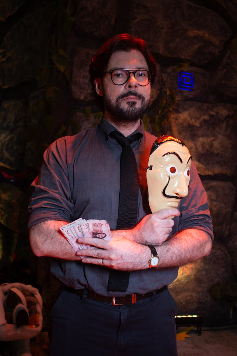 A bearded man stands in a dimly lit room holding a mask and money, exuding an air of mystery.