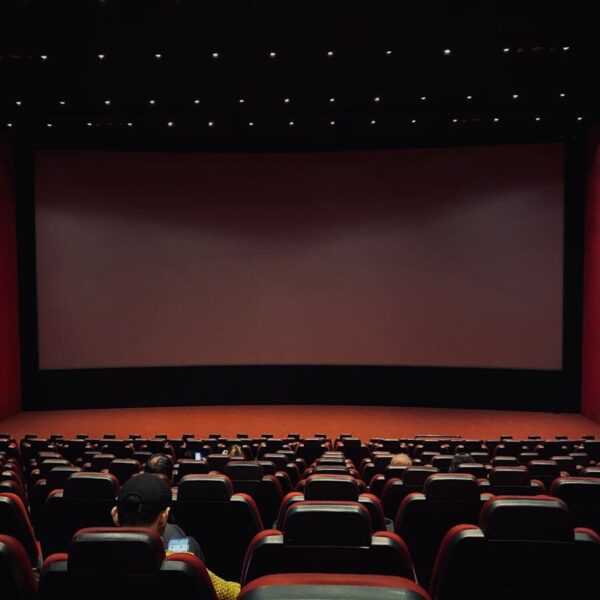 Dimly lit cinema hall with red seats and a blank screen. Minimal audience.