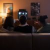 Three friends sitting on a couch enjoy a cozy movie night together, pointing at the TV screen.