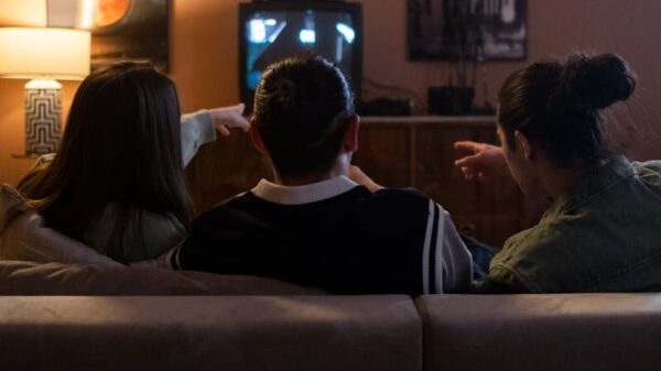 Three friends sitting on a couch enjoy a cozy movie night together, pointing at the TV screen.