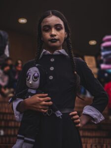 A young girl dressed in a dark costume holds a gothic doll, capturing a mysterious and dramatic vibe.