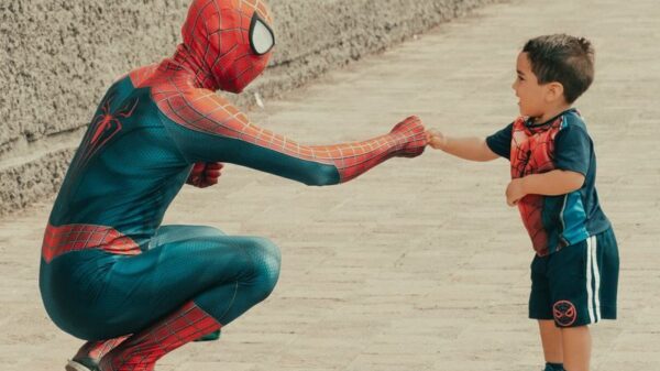 A child dressed in superhero attire shares a fist bump with a man in a Spiderman costume outdoors.