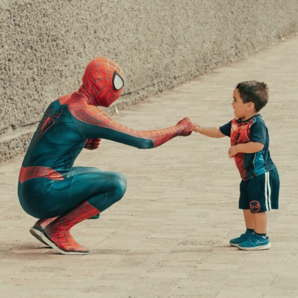 A child dressed in superhero attire shares a fist bump with a man in a Spiderman costume outdoors.