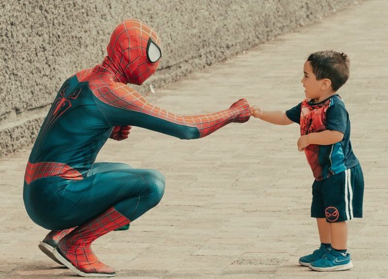 A child dressed in superhero attire shares a fist bump with a man in a Spiderman costume outdoors.