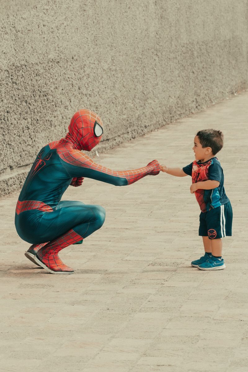 A child dressed in superhero attire shares a fist bump with a man in a Spiderman costume outdoors.