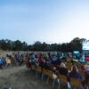 A large group enjoys a movie outdoors at night in a field with a big screen under the moon.