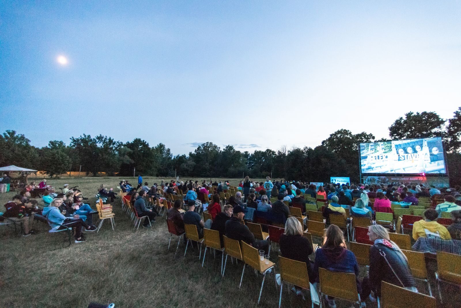 A large group enjoys a movie outdoors at night in a field with a big screen under the moon.