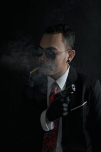 A man in a suit and sunglasses holding a gun while smoking in a dark studio setting.