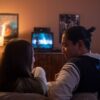 A couple enjoys a cozy movie night at home, watching TV on a couch in dim lighting.