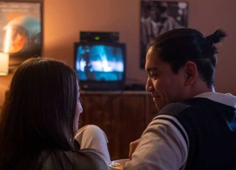 A couple enjoys a cozy movie night at home, watching TV on a couch in dim lighting.
