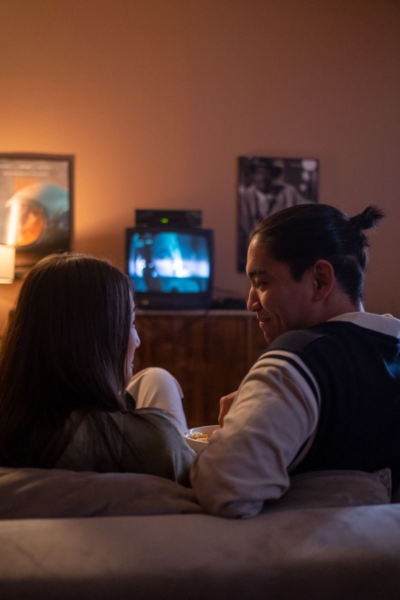 A couple enjoys a cozy movie night at home, watching TV on a couch in dim lighting.