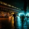A pedestrian walks on a wet urban street under an illuminated train at night, capturing the moody city atmosphere.