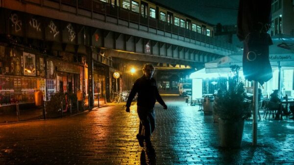 A pedestrian walks on a wet urban street under an illuminated train at night, capturing the moody city atmosphere.