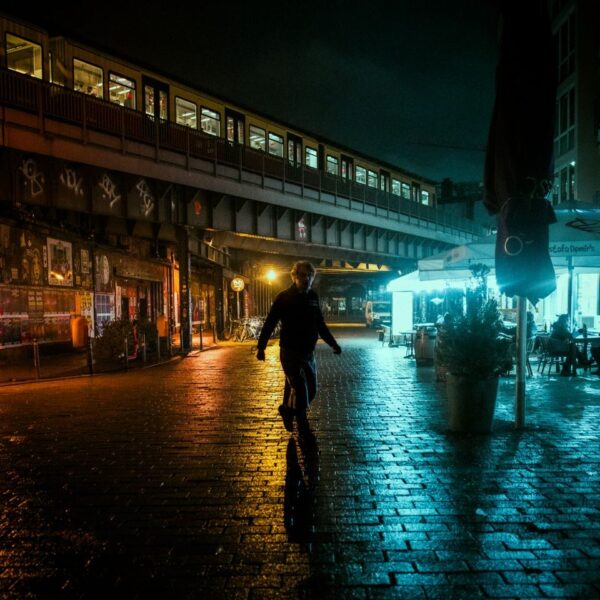 A pedestrian walks on a wet urban street under an illuminated train at night, capturing the moody city atmosphere.