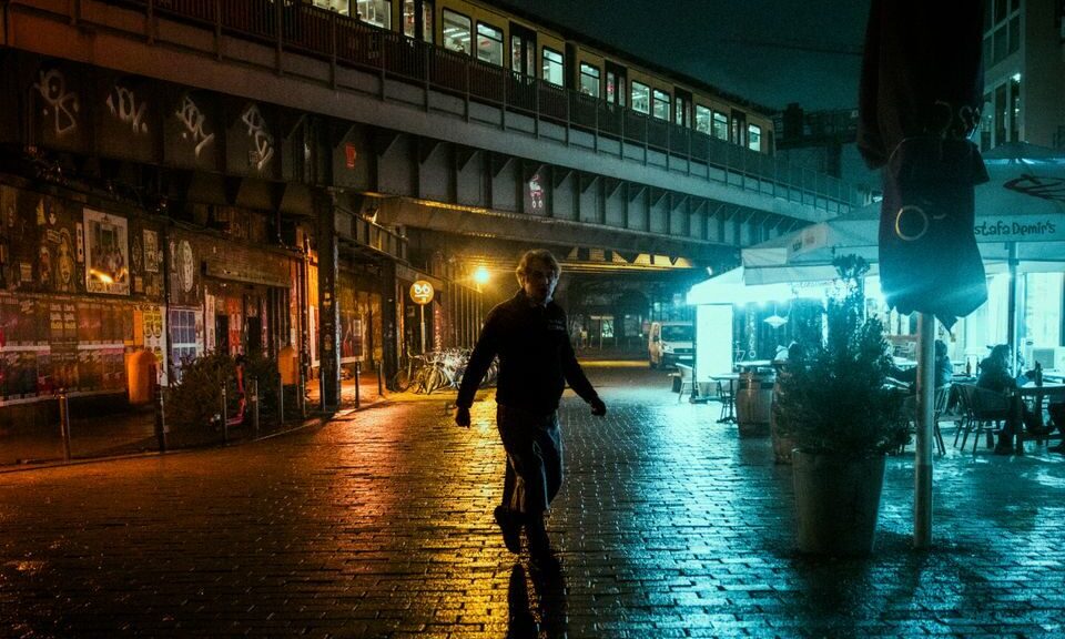 A pedestrian walks on a wet urban street under an illuminated train at night, capturing the moody city atmosphere.