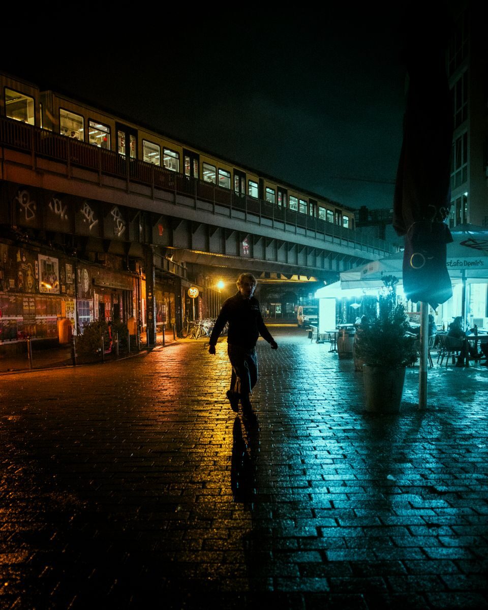 A pedestrian walks on a wet urban street under an illuminated train at night, capturing the moody city atmosphere.