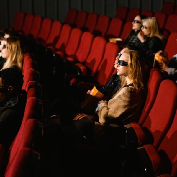 People wearing 3D glasses and eating popcorn in a cinema with red chairs.