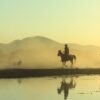 Silhouetted horse riders at sunset with misty mountain reflection in the lake.