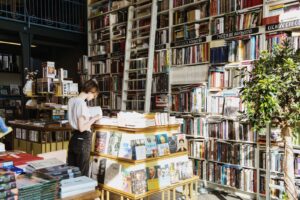 A person reads a book in a cozy, sunlit bookstore filled with shelves of books.