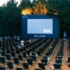 An outdoor cinema setup with numerous empty chairs and a large screen surrounded by trees.