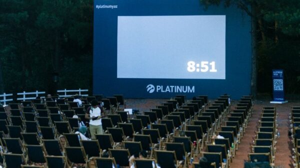 An outdoor cinema setup with numerous empty chairs and a large screen surrounded by trees.