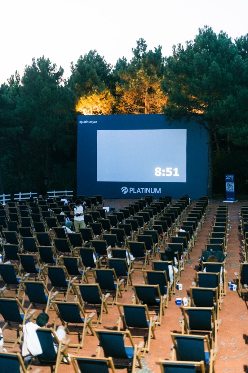 An outdoor cinema setup with numerous empty chairs and a large screen surrounded by trees.