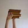 Hands reaching upwards holding a stack of hardbound books against a plain background.