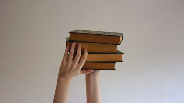 Hands reaching upwards holding a stack of hardbound books against a plain background.