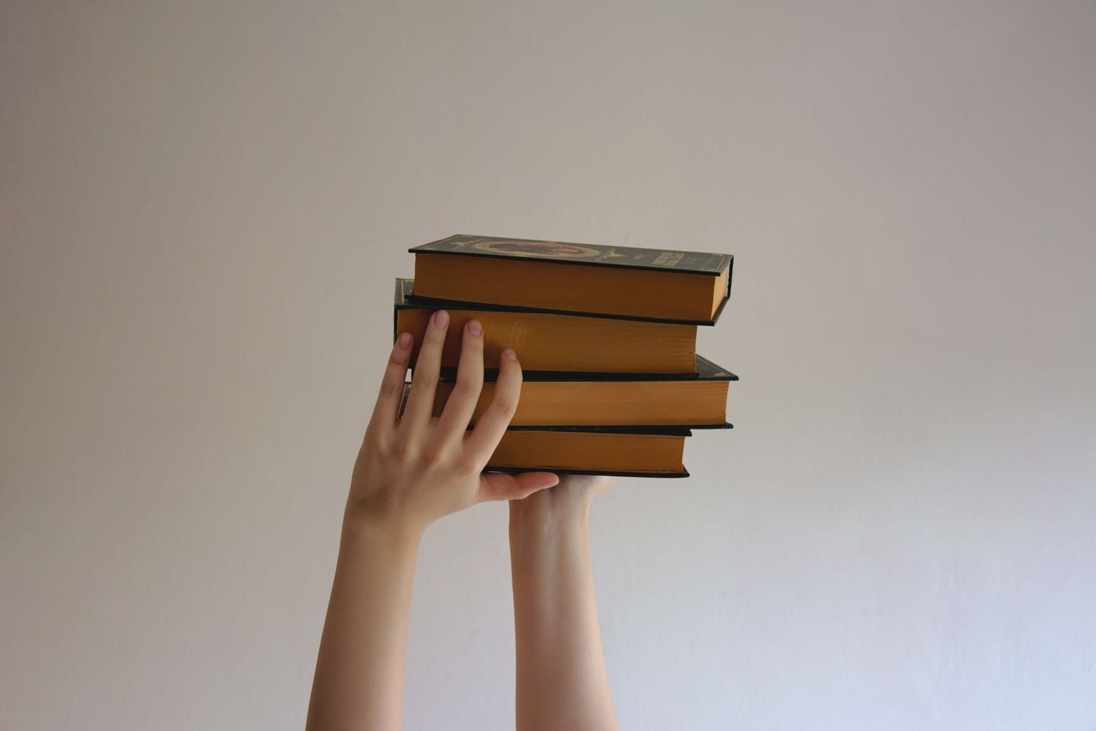 Hands reaching upwards holding a stack of hardbound books against a plain background.