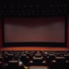 Dimly lit cinema hall with red seats and a blank screen. Minimal audience.