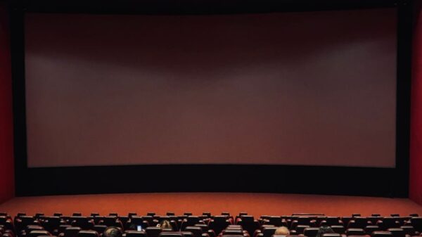 Dimly lit cinema hall with red seats and a blank screen. Minimal audience.