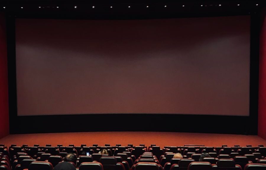 Dimly lit cinema hall with red seats and a blank screen. Minimal audience.