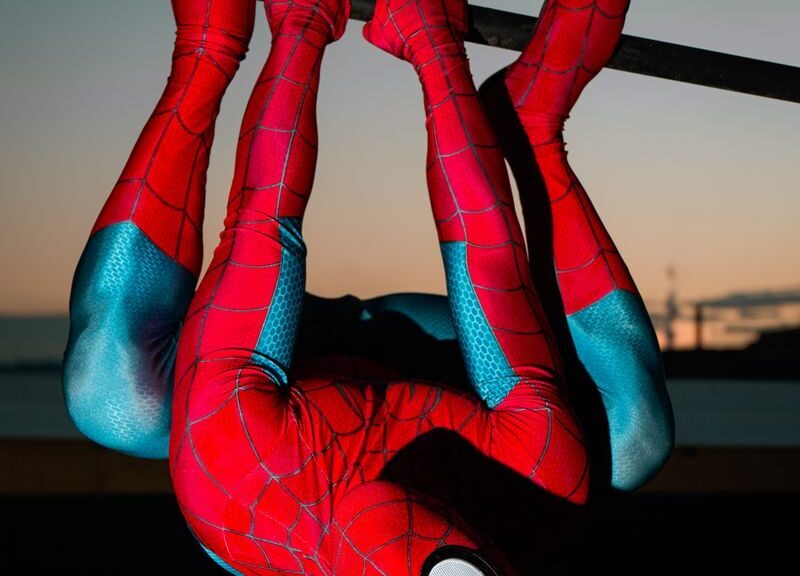 A superhero in red and blue costume hangs upside down on bars at sunset.