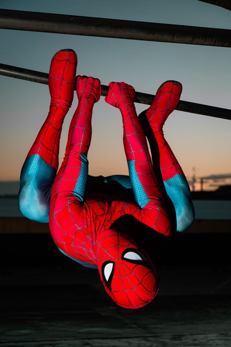 A superhero in red and blue costume hangs upside down on bars at sunset.