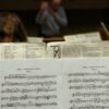 Detailed shot of sheet music during a symphony rehearsal, conductor in the background.