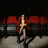 A stylish woman sits alone in a dark cinema, enjoying popcorn during a movie.