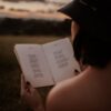 Back view of anonymous female in hat reading book with poetry in grassy field on blurred background on summer evening
